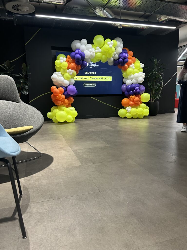 A bold and playful organic balloon arch created for the London College of Contemporary Arts, featuring an energetic mix of neon yellow, orange, purple and white balloons in varying sizes for a modern, high-impact finish.