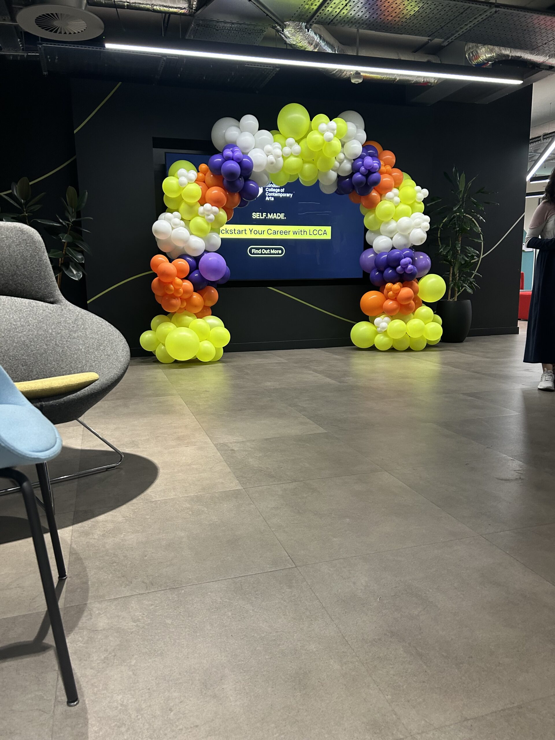 A bold and playful organic balloon arch created for the London College of Contemporary Arts, featuring an energetic mix of neon yellow, orange, purple and white balloons in varying sizes for a modern, high-impact finish.